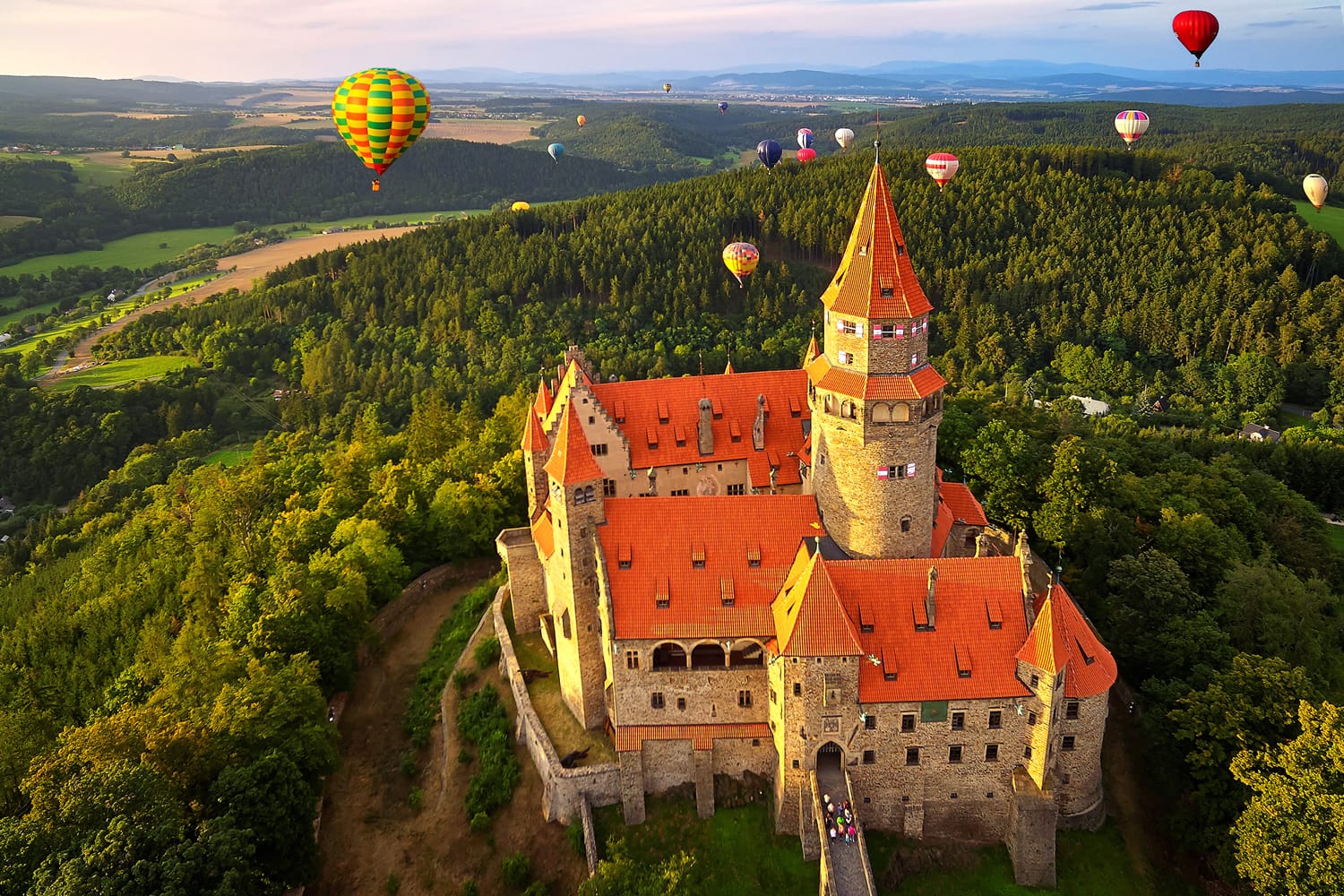 Aerial view on romantic fairy castle with group hot air balloons in picturesque landscape lit by evening sun. Moravia, Czech republic.