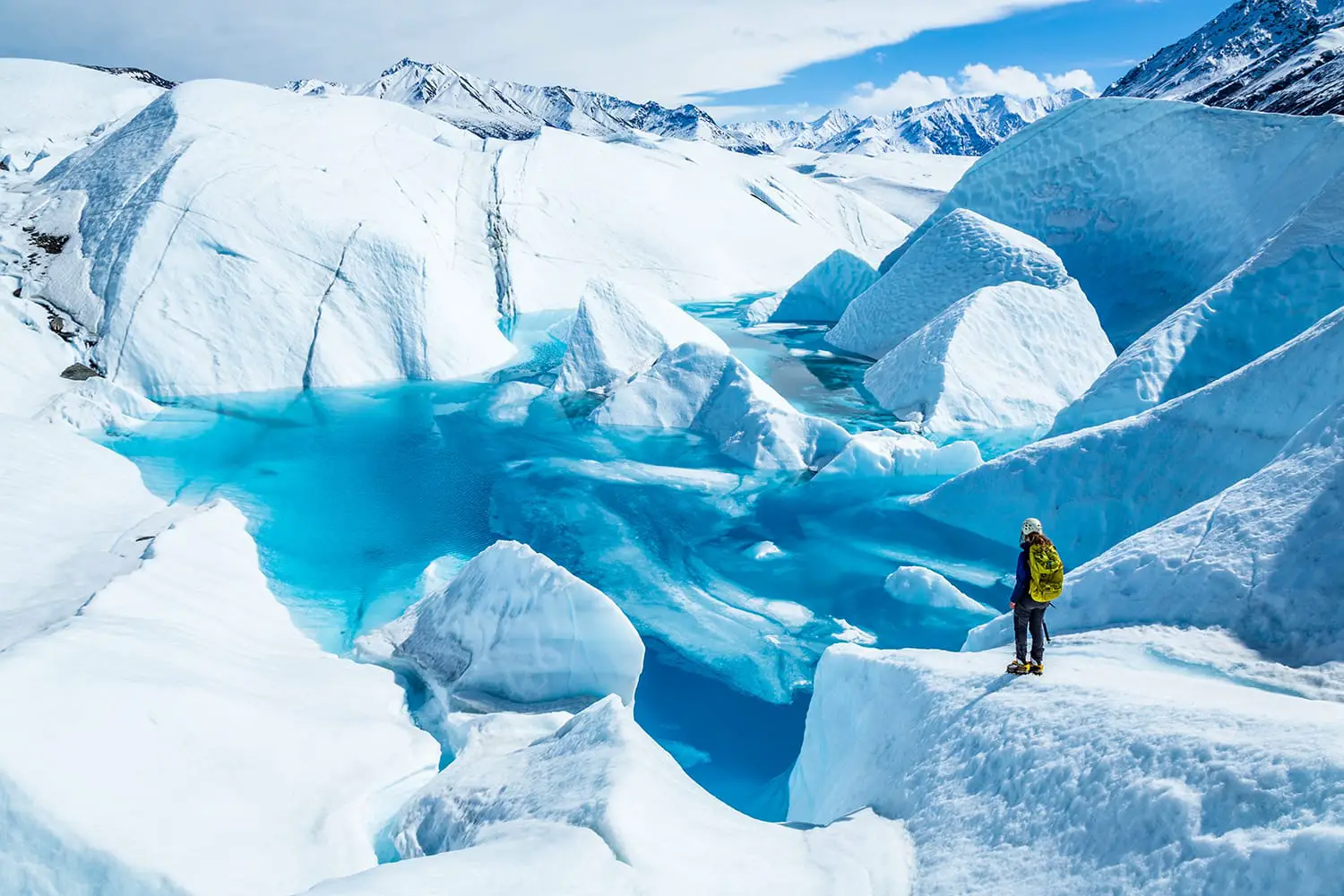 Standing near the edge of large blue pool on top of the Matanuska Glacier. A young woman holding an ice axe with a backpack and helmet looks out over the lake.