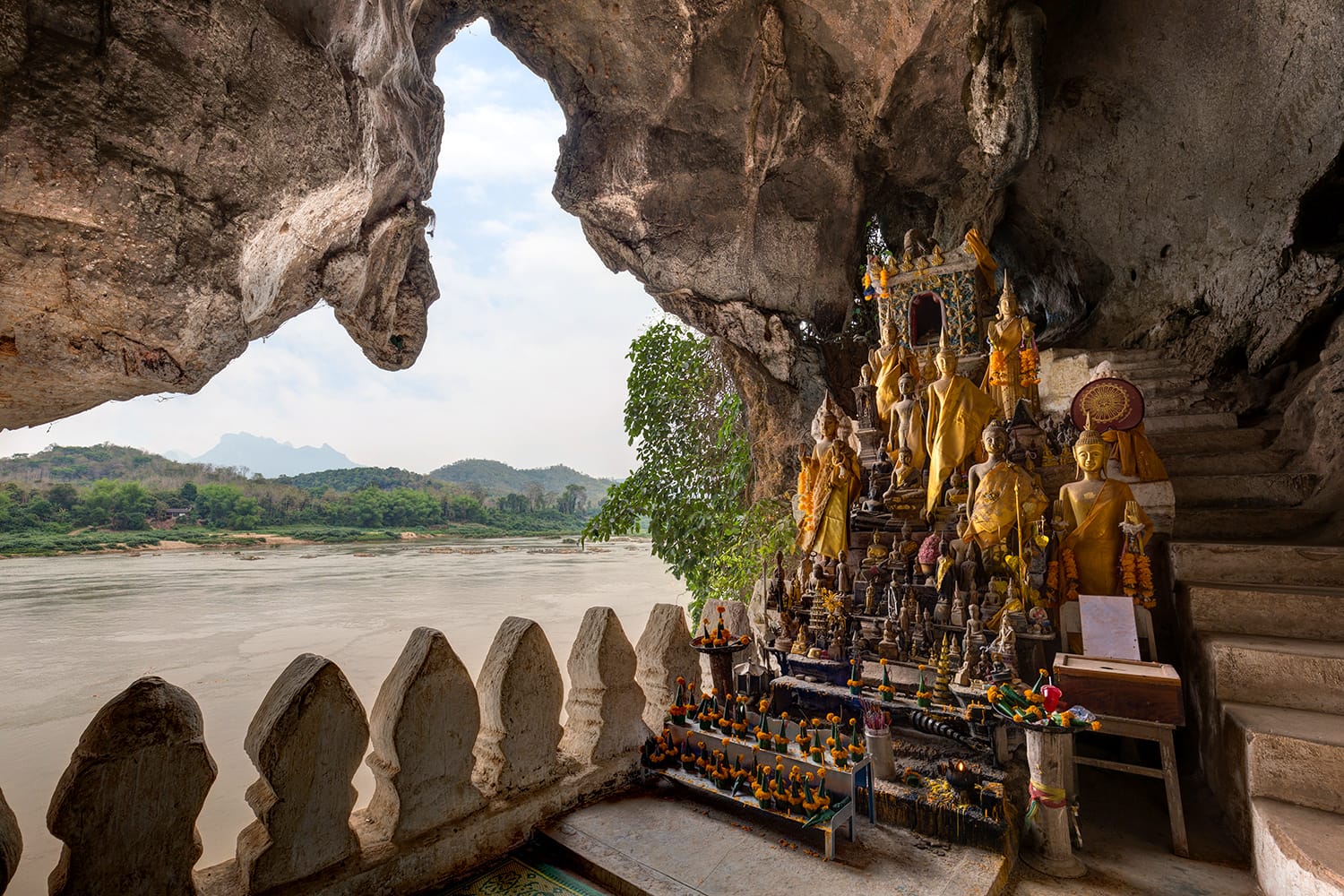 View of the Mekong River and many golden and wooden Buddha statues and religious offerings inside the Tham Ting Cave at the famous Pak Ou Caves near Luang Prabang in Laos.