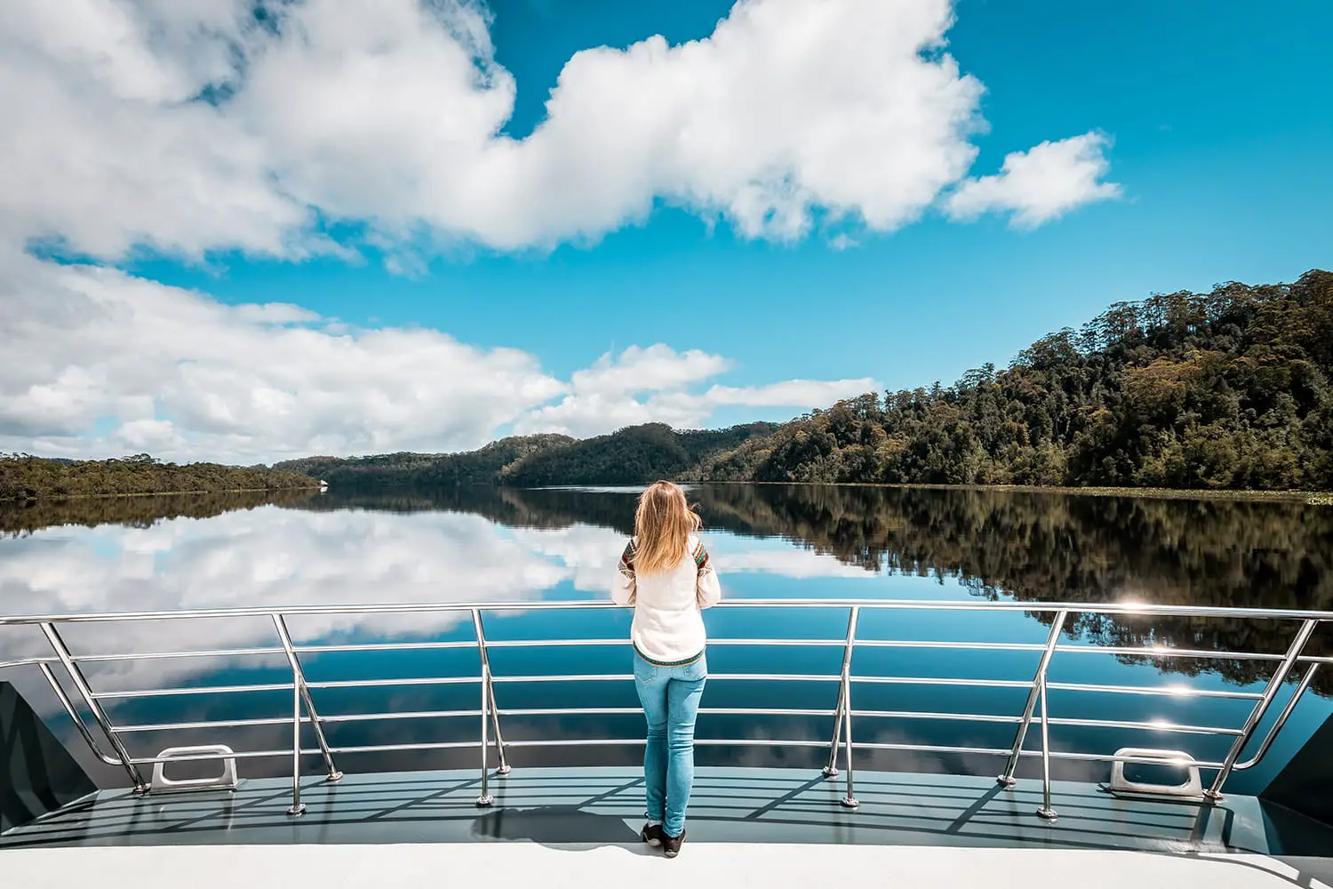 Woman stands by the railing of a ship and looks at the mirror-like water, surrounded by wilderness, sunny blue sky, Gordon River, Tasmania, Australia
